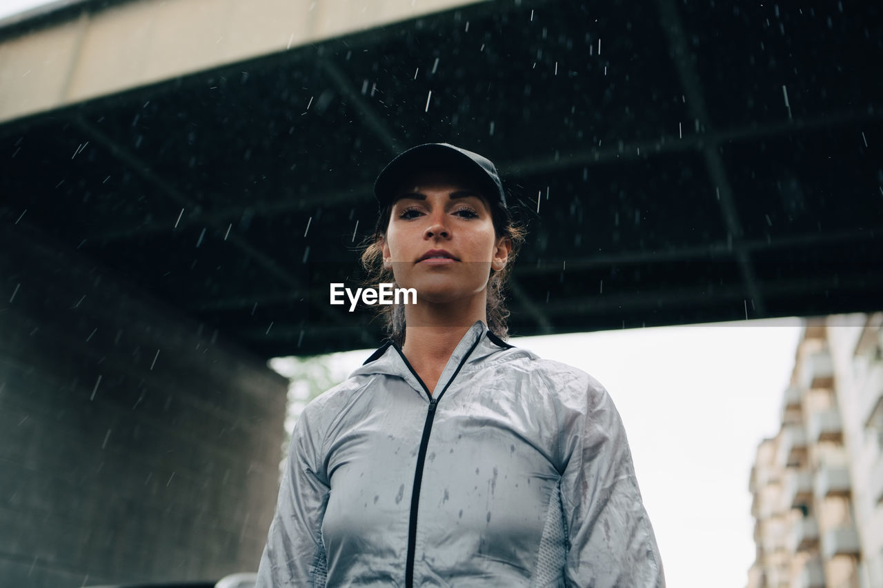 Low angle portrait of female athlete standing against bridge during rainy season