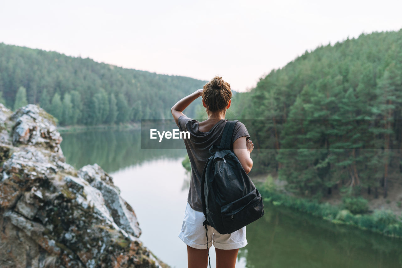 Young slim woman in casual clothes with backpack looks at beautiful view of mountains and river