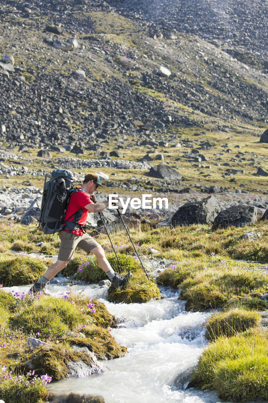 Backpacker steps across river in alpine meadow.