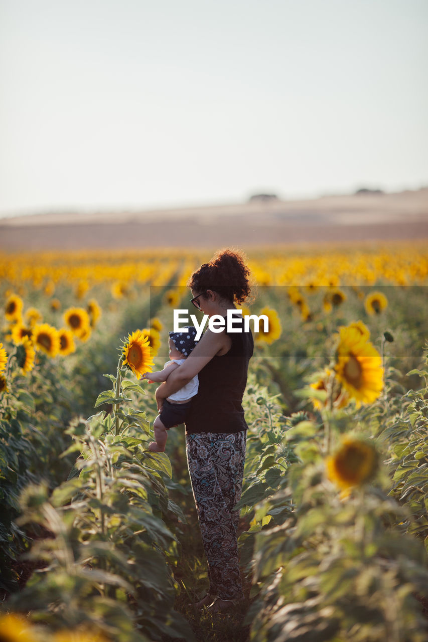 Side view of mother carrying daughter while standing amidst sunflowers in field against sky