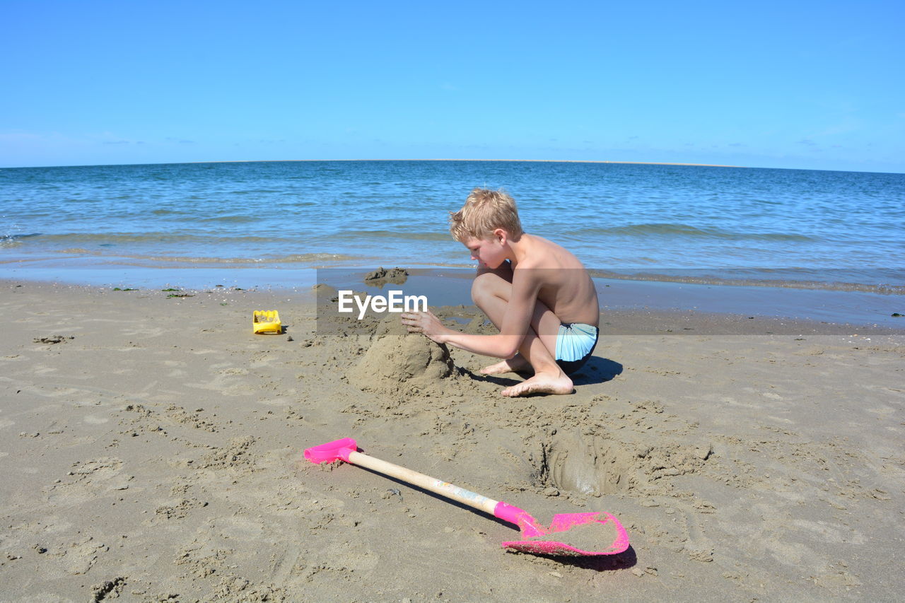 Full length side view of shirtless boy playing with sand at beach against sky during summer