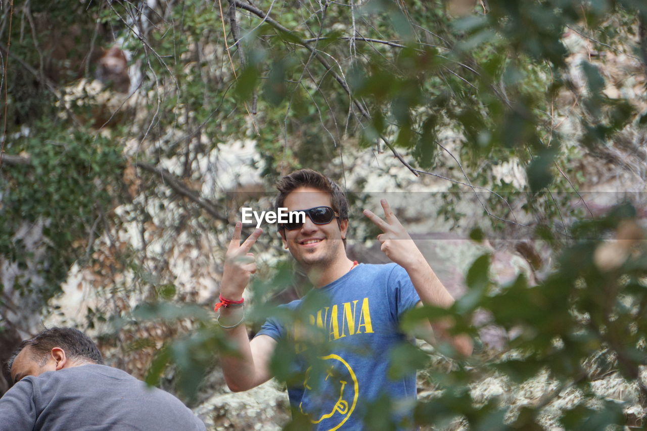 Portrait of man in sunglasses gesturing peace signs amidst trees in forest