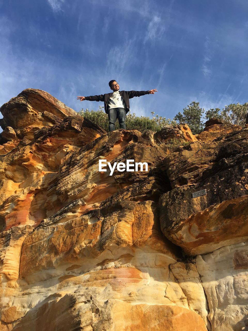 Low angle view of young man with arms outstretched standing on rock against blue sky during sunny day