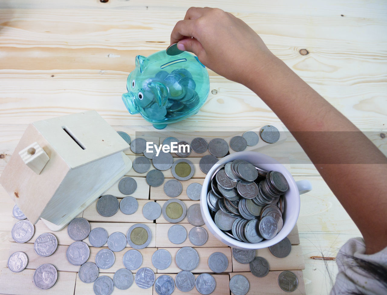 Person holding coin over piggy bank by wooden model home and cup on table