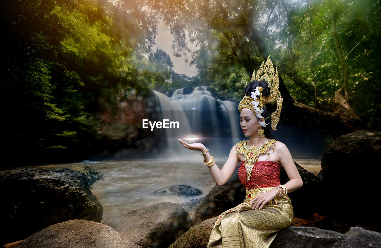 Beautiful young woman wearing crown and traditional clothing while sitting against waterfall in forest