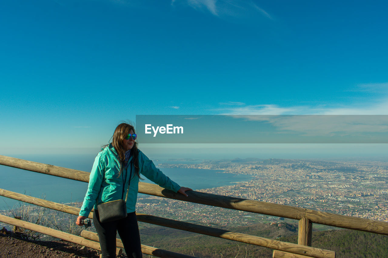 Mid adult woman standing by railing on observation point against blue sky during sunny day