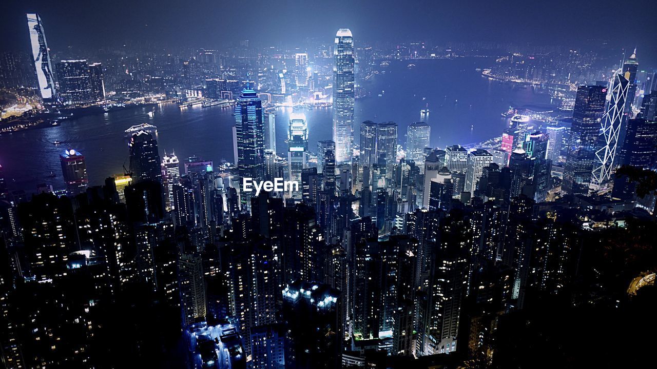 Illuminated cityscape by bay against sky
