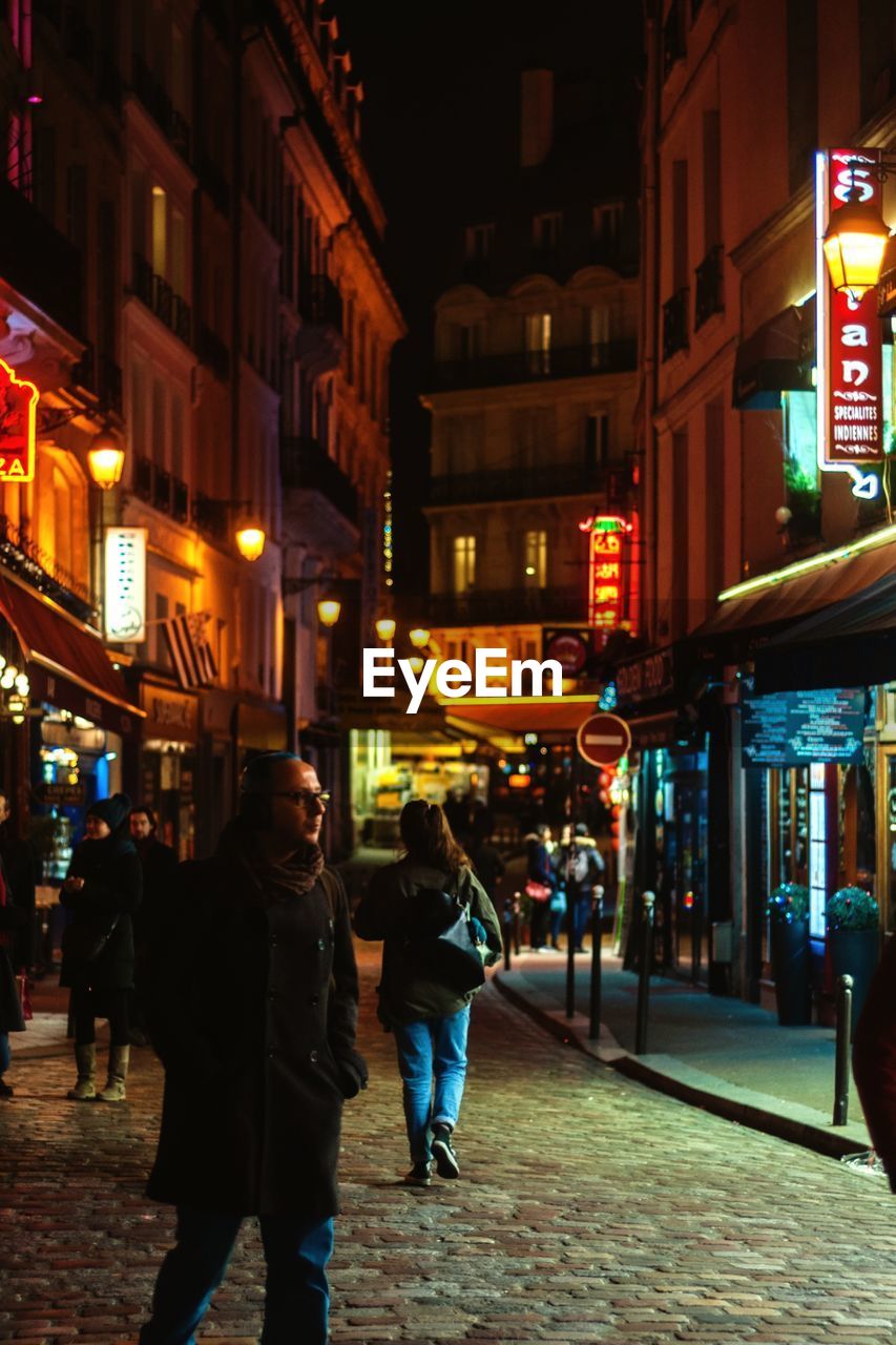 PEOPLE WALKING ON ILLUMINATED STREET AMIDST BUILDINGS