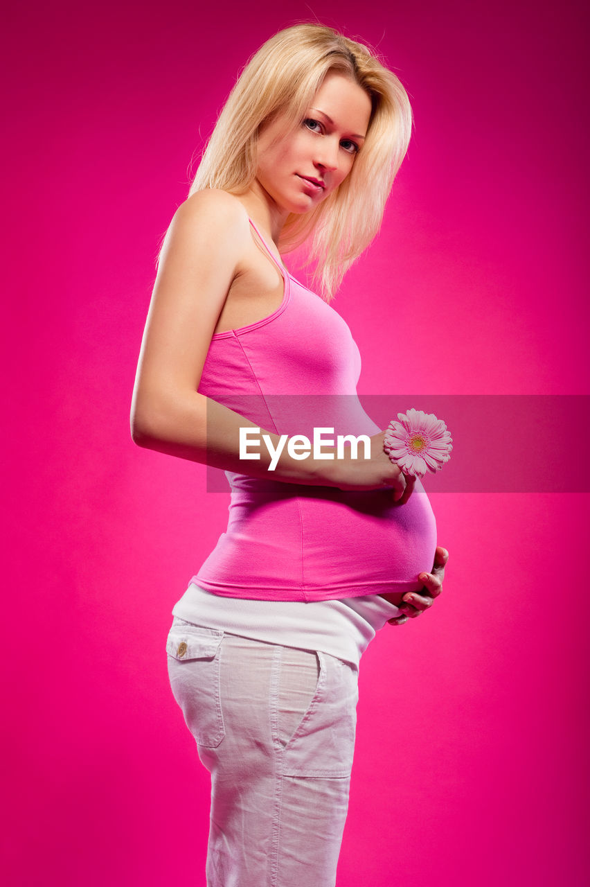 Portrait of pregnant woman holding flower standing against pink background