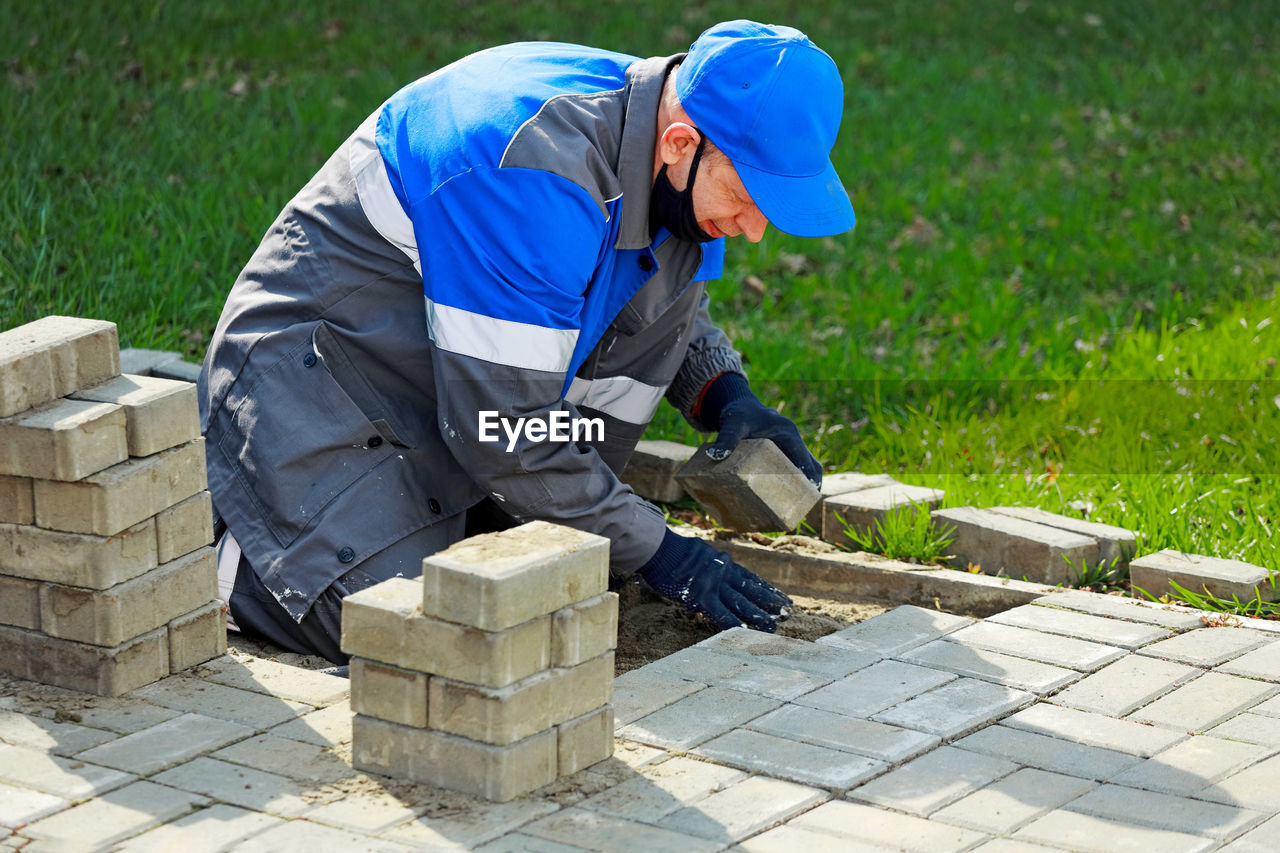 Bricklayer in work clothes sits on sidewalk and lays out paving slabs. sight of working man in open