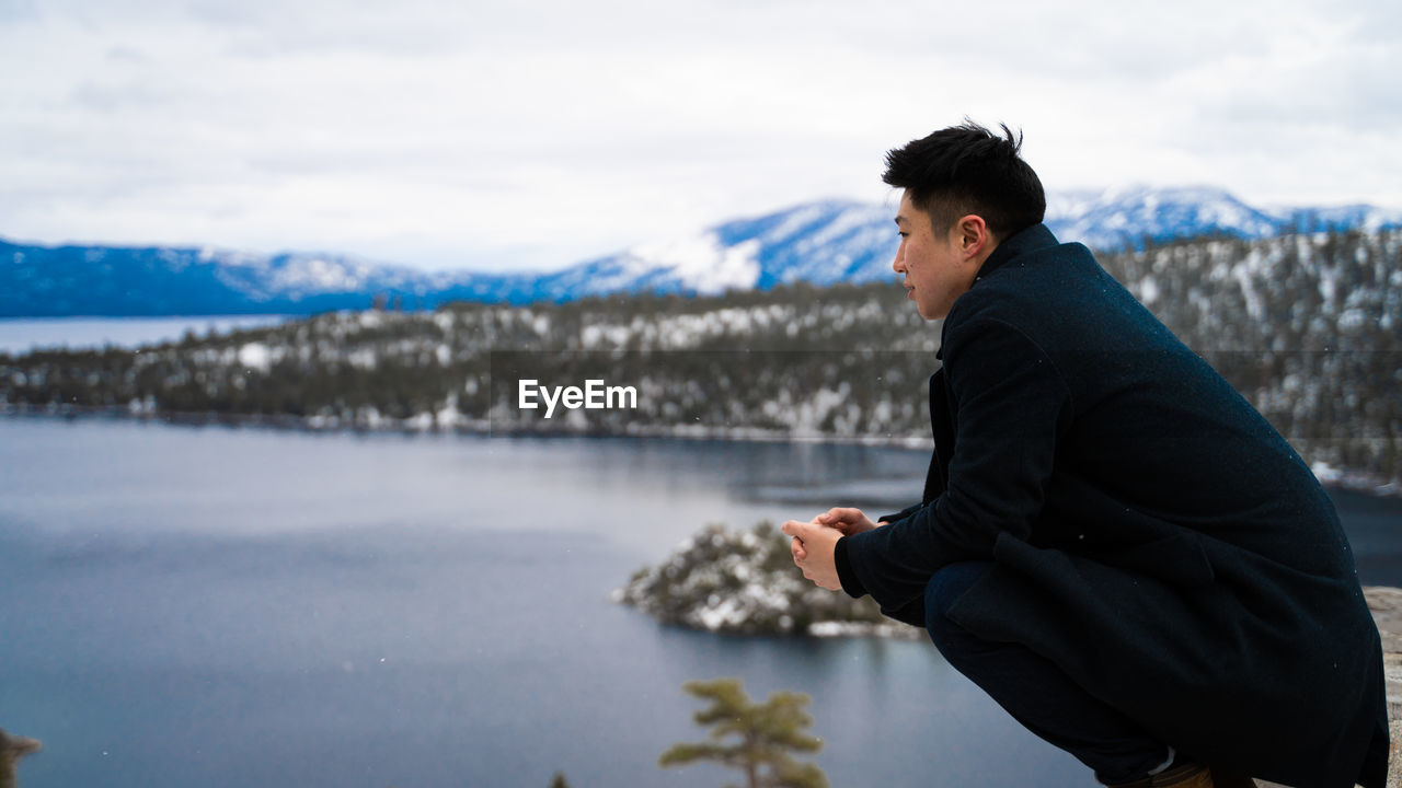 Side view of young man crouching over lake against sky