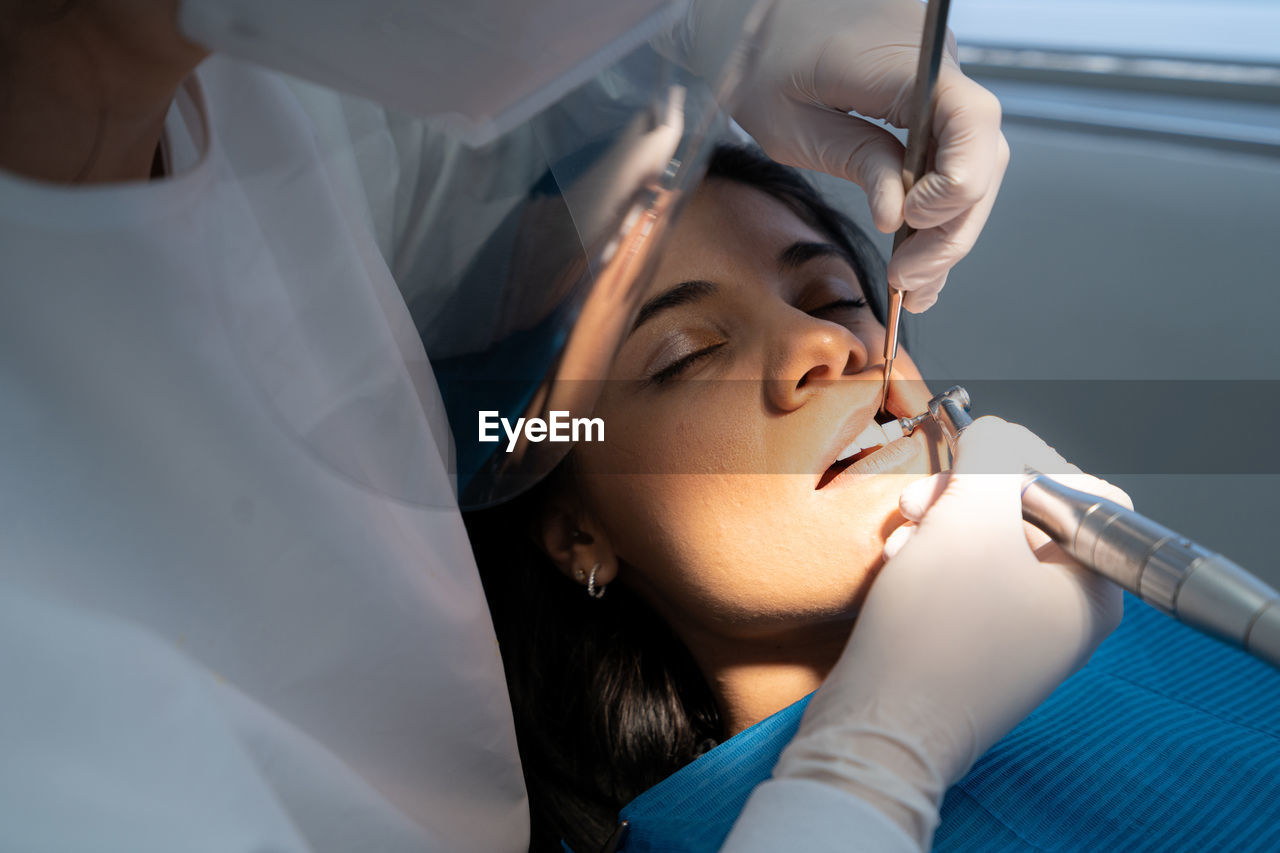 Crop professional dentist in uniform with medical mask drilling tooth of calm woman with help of assistant