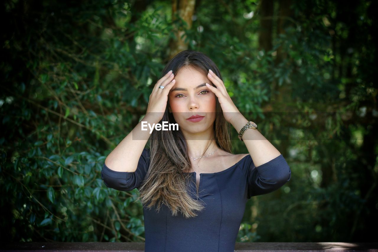 Portrait of beautiful young woman standing against plants seriously touching the hair