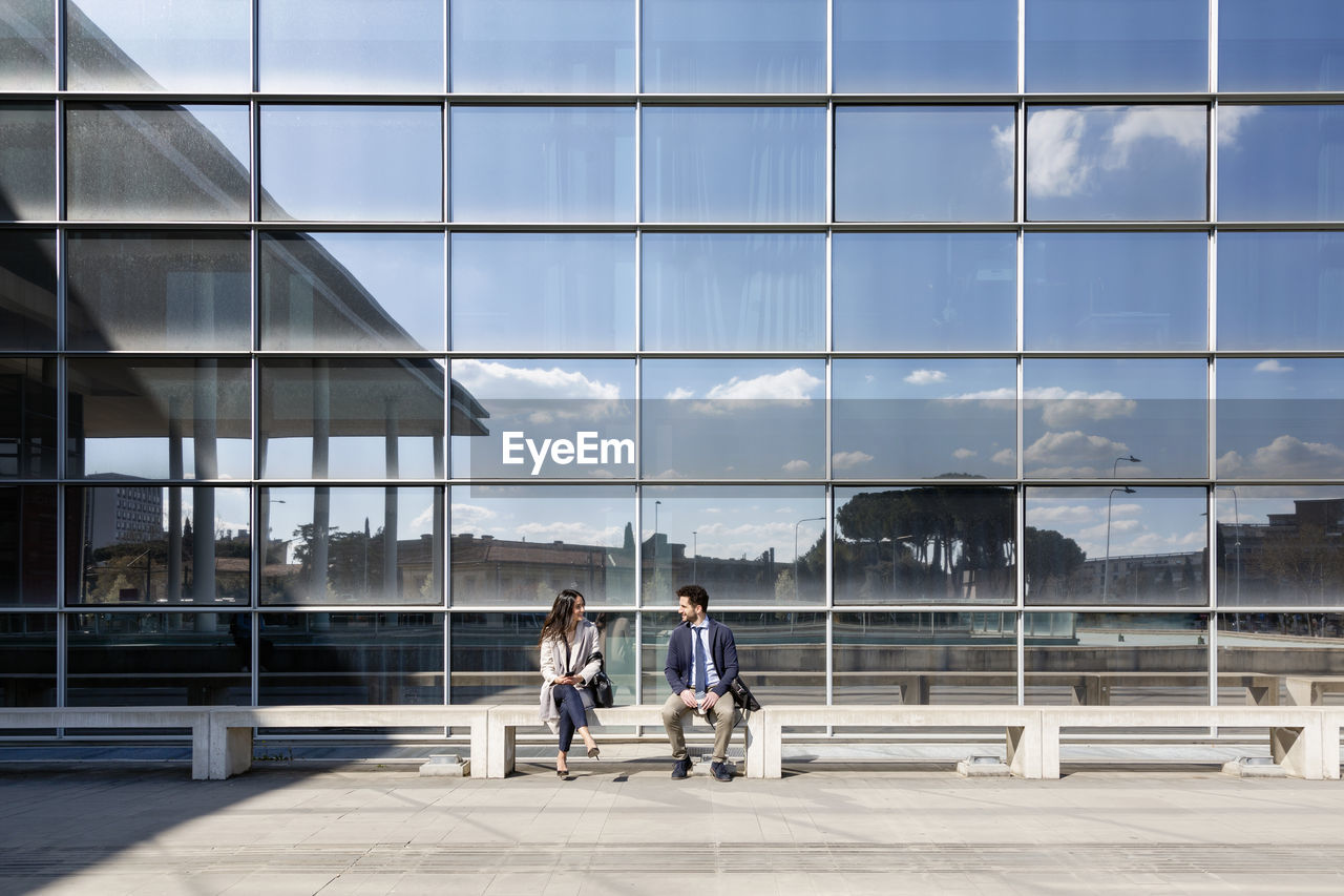 Business couple talking while sitting on bench during sunny day