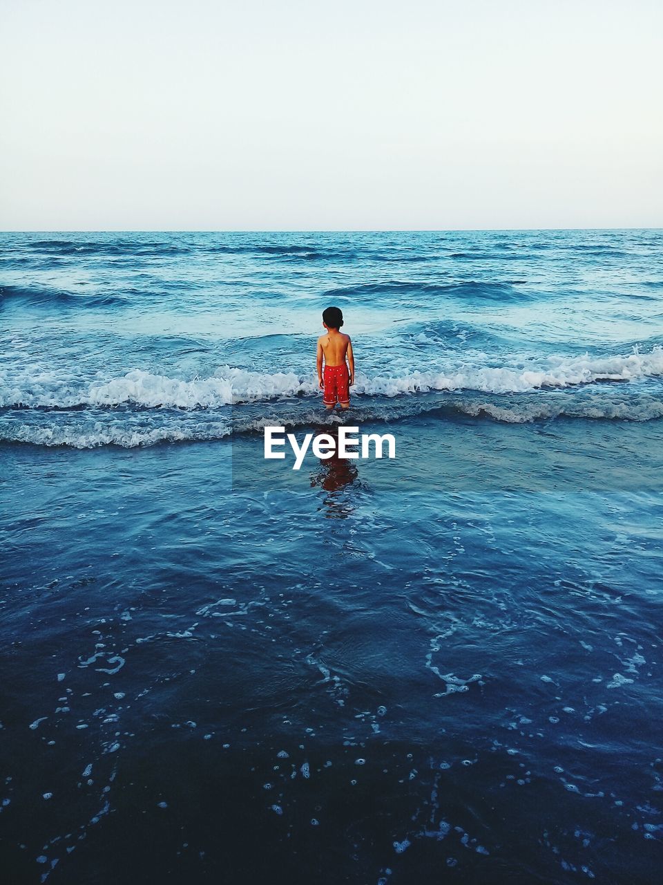 Rear view of shirtless boy standing in sea on shore against clear sky