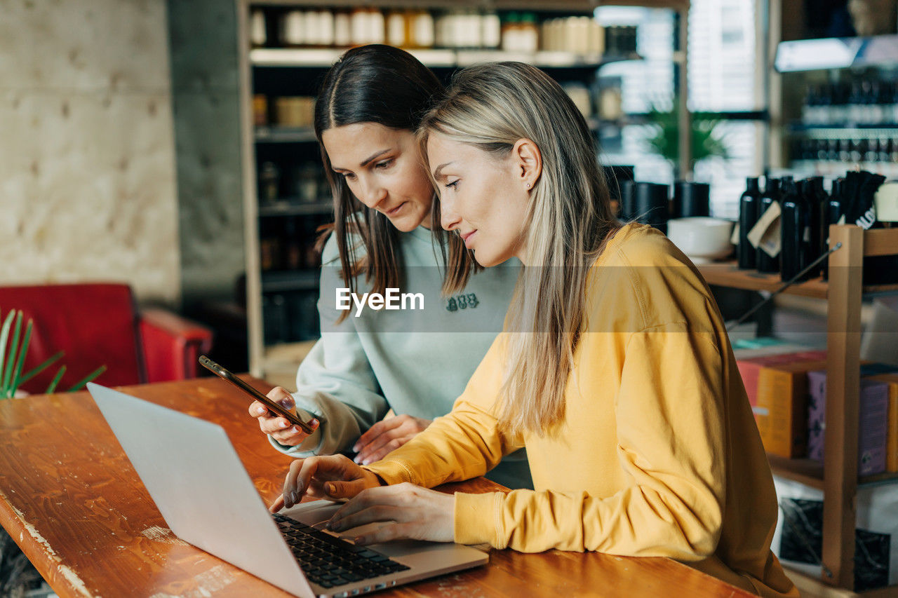 Two business women work behind the counter using electronic gadgets.