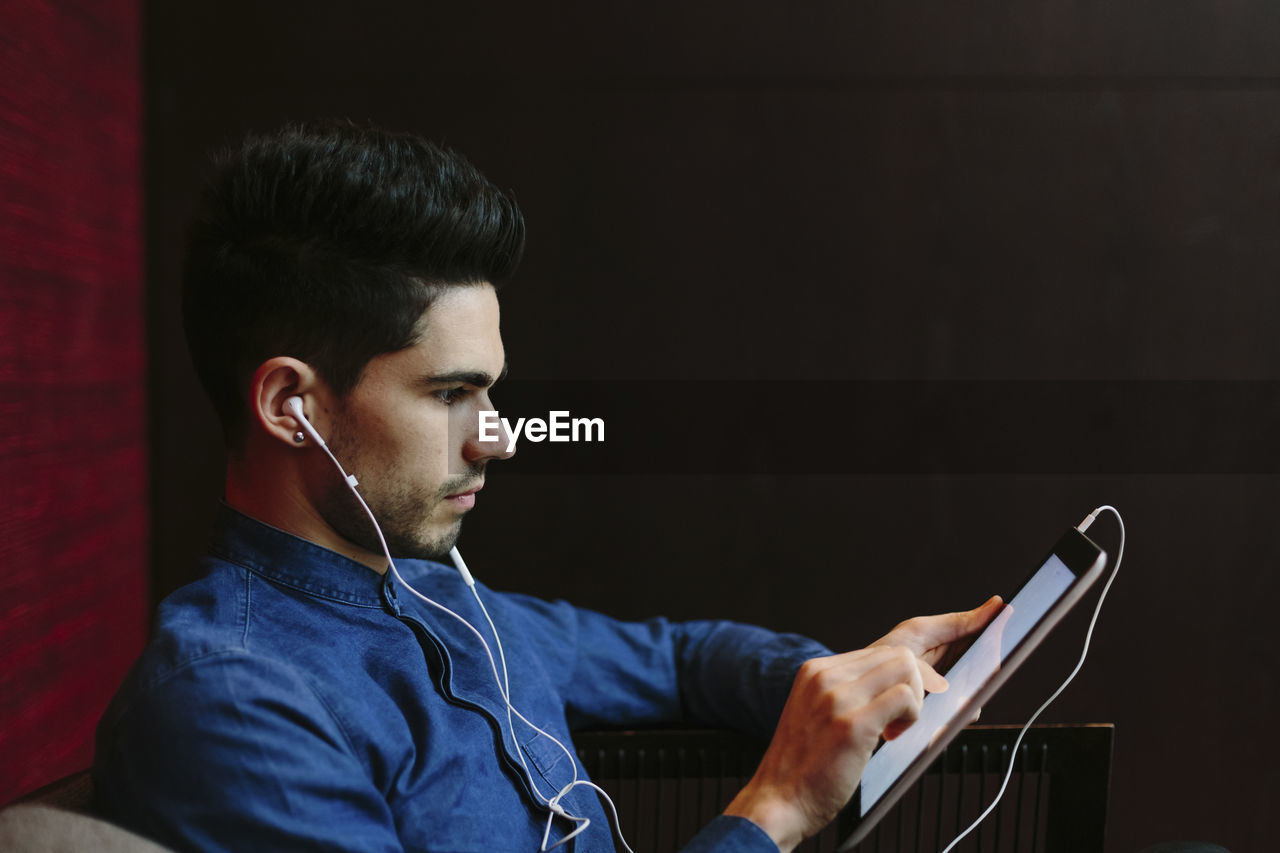 Profile of young businessman with earphones using digital tablet in front of black background