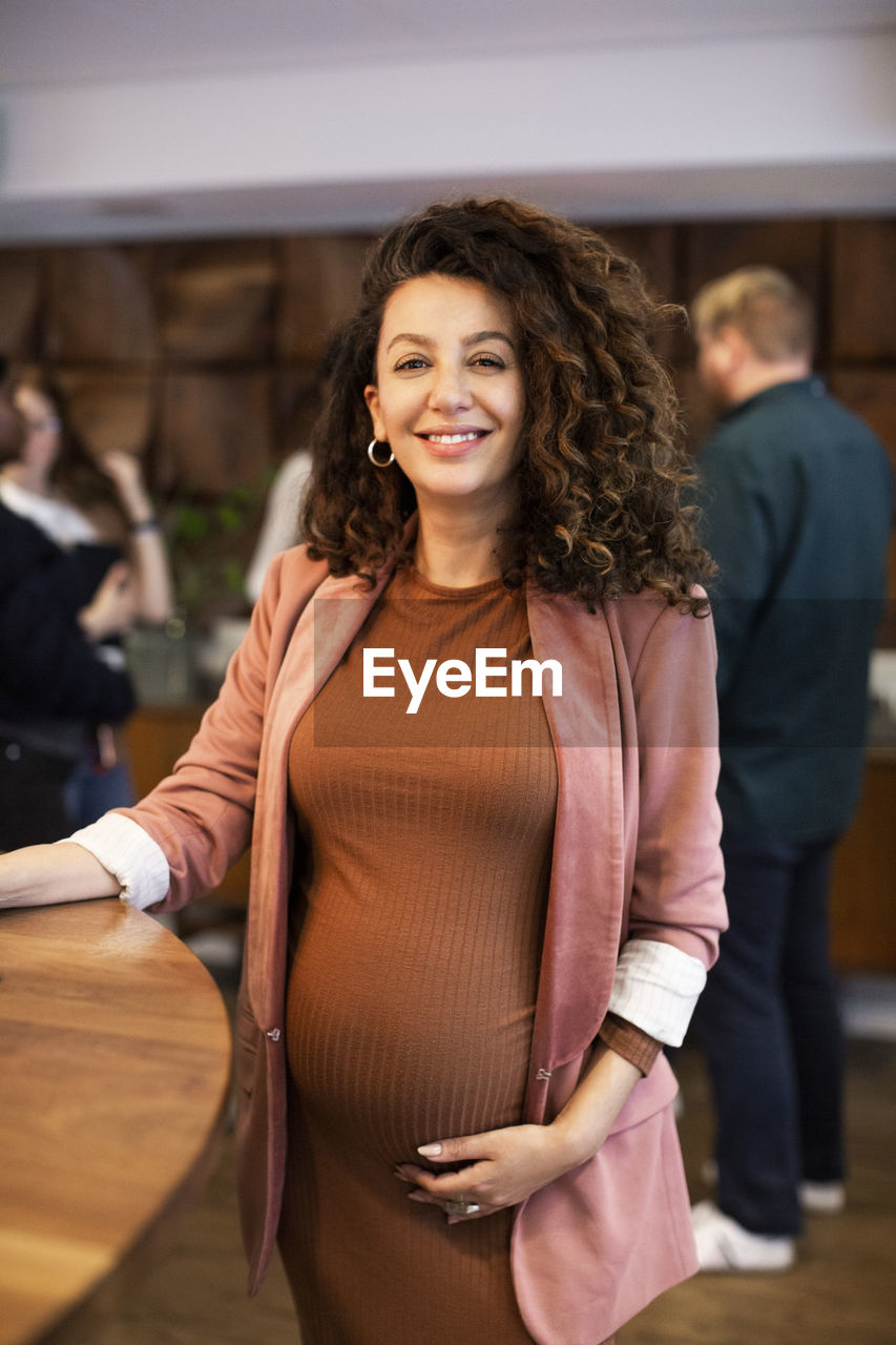 Portrait of pregnant female boss standing by desk in office