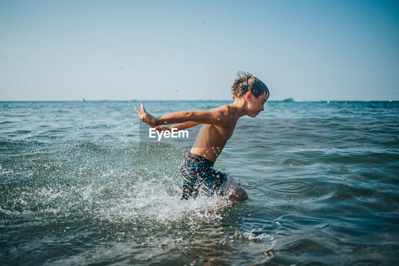 Shirtless boy playing in water against sky