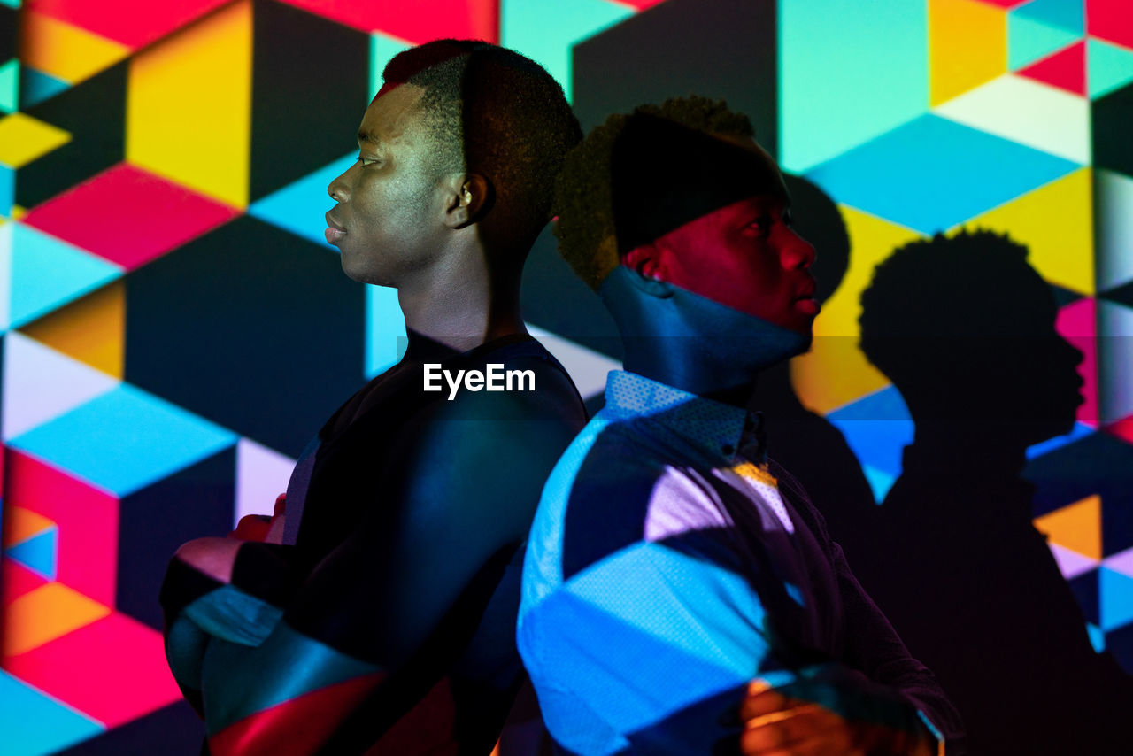 Side view of emotionless african american men standing back to back in studio with colorful neon illumination in shape of cubes