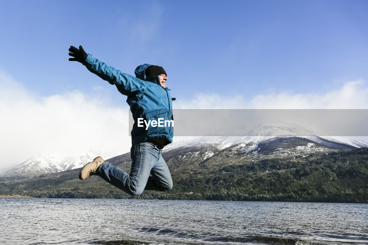 Excited hiker jumping by river against sky