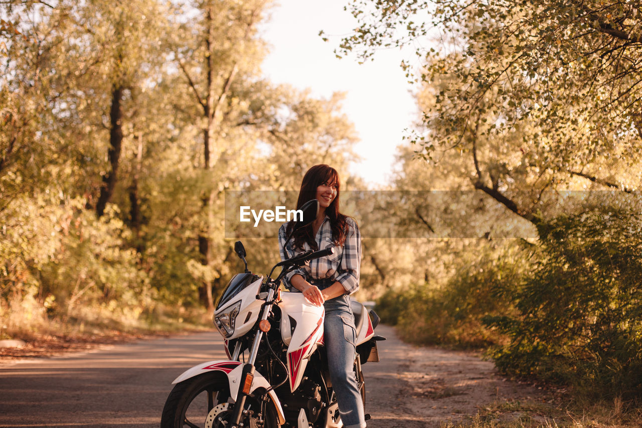 Smiling young woman sitting on motorcycle on country road amidst trees