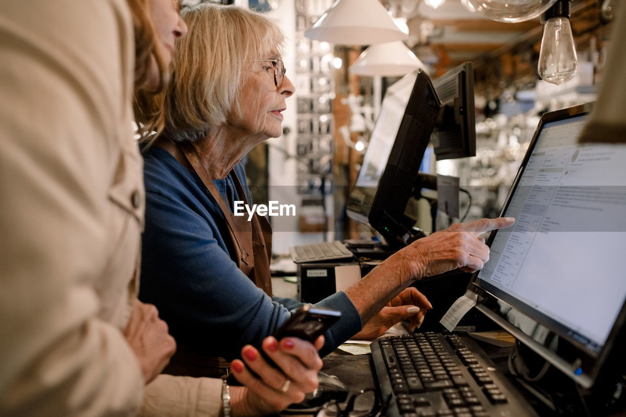 Senior female customer and saleswoman discussing over computer monitor at checkout in hardware store