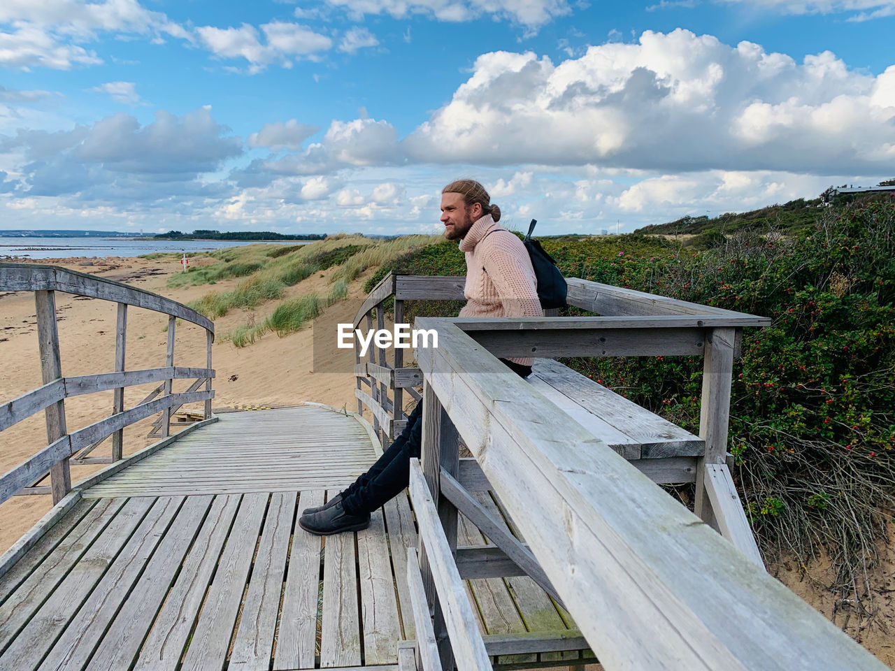 Man sitting on wooden railing against sky