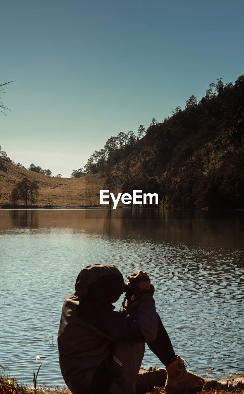 Man photographing while sitting by lake against clear sky