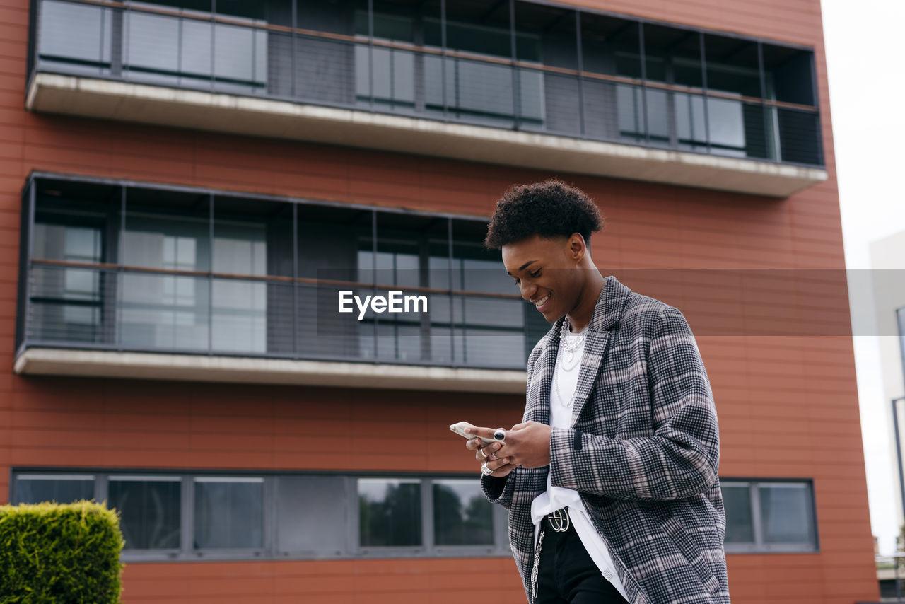 From below side view of happy young african american male student in stylish outfit browsing smartphone while standing on city street near modern building