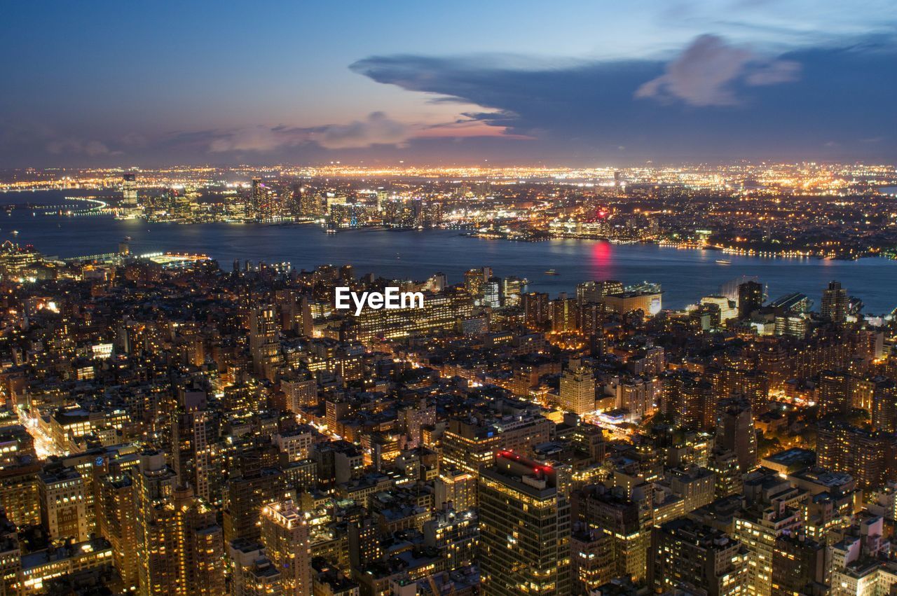 High angle view of illuminated cityscape by river against cloudy sky at dusk