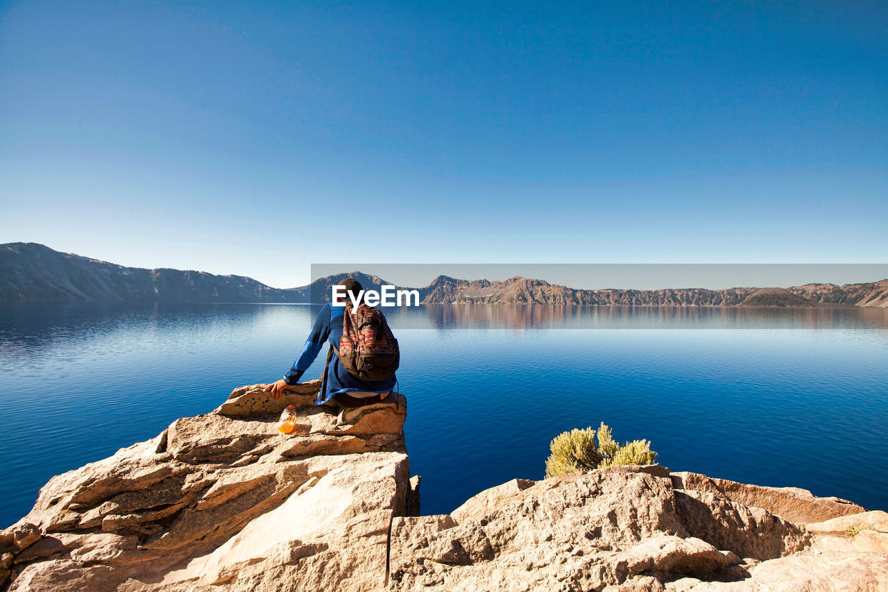 Man sitting on rock at lake against clear blue sky