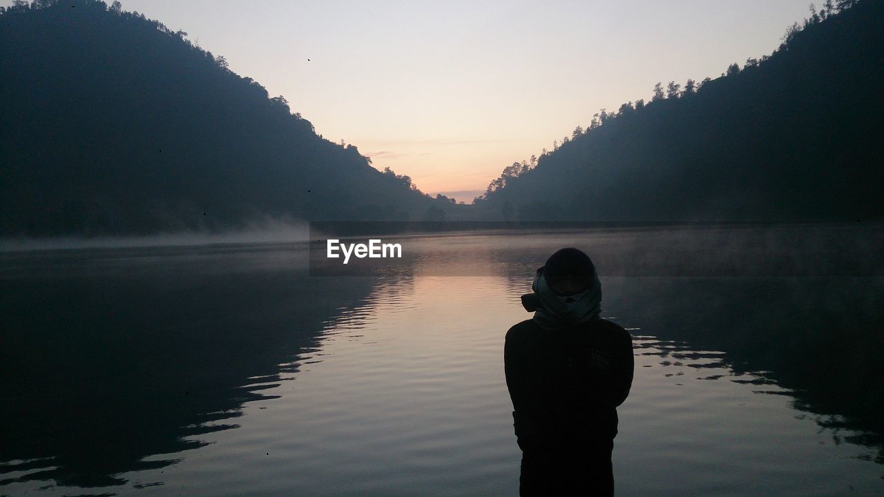 Rear view of man standing by lake against sky during sunset