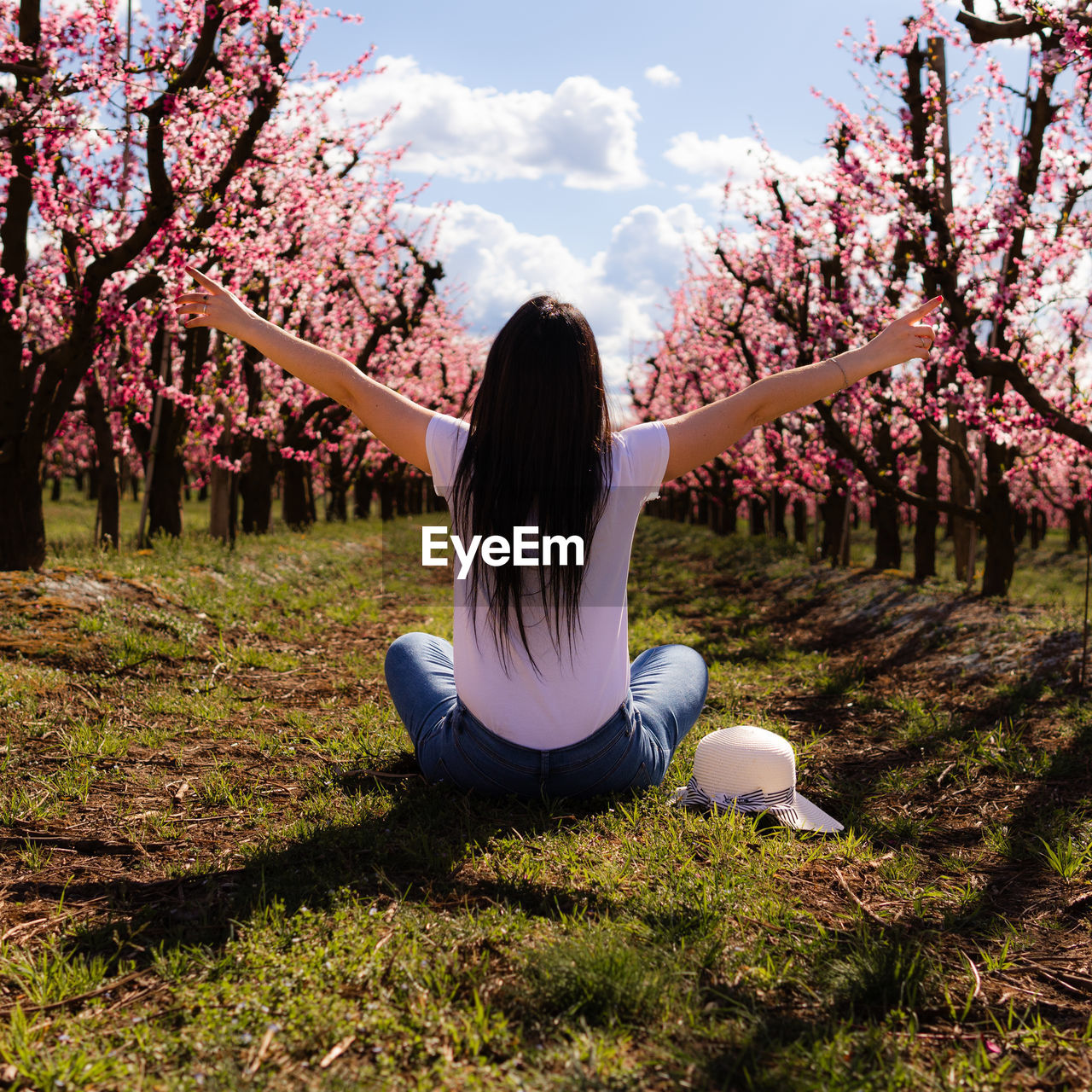 Unrecognizable woman sitting on her back with open arms in a field of peach trees with flowers