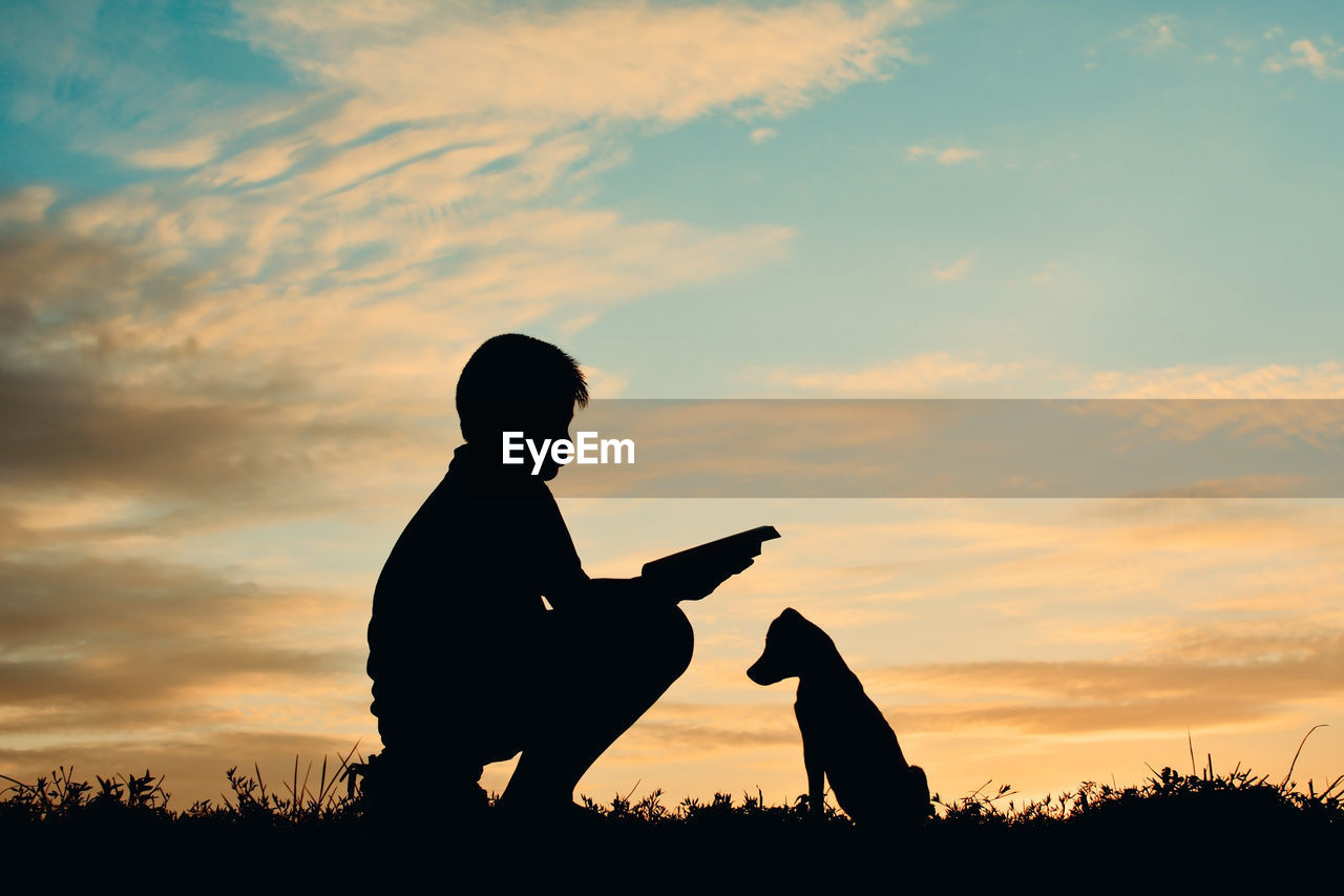 Silhouette boy reading book while sitting by puppy against cloudy sky during sunset