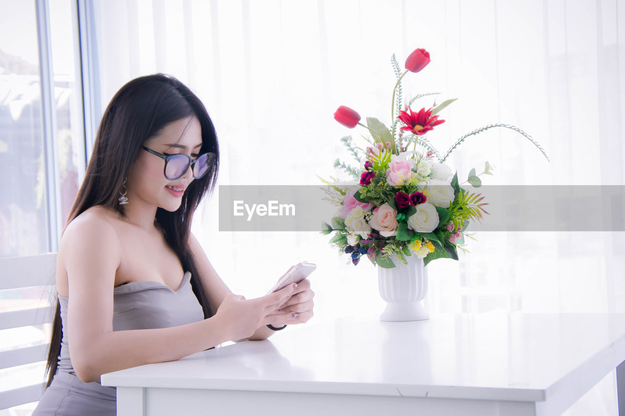 Woman using mobile phone by flower vase at home