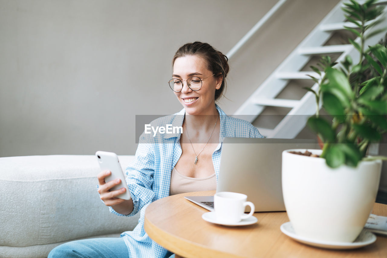 Smiling brunette woman with long hair working on laptop using mobile phone in bright home