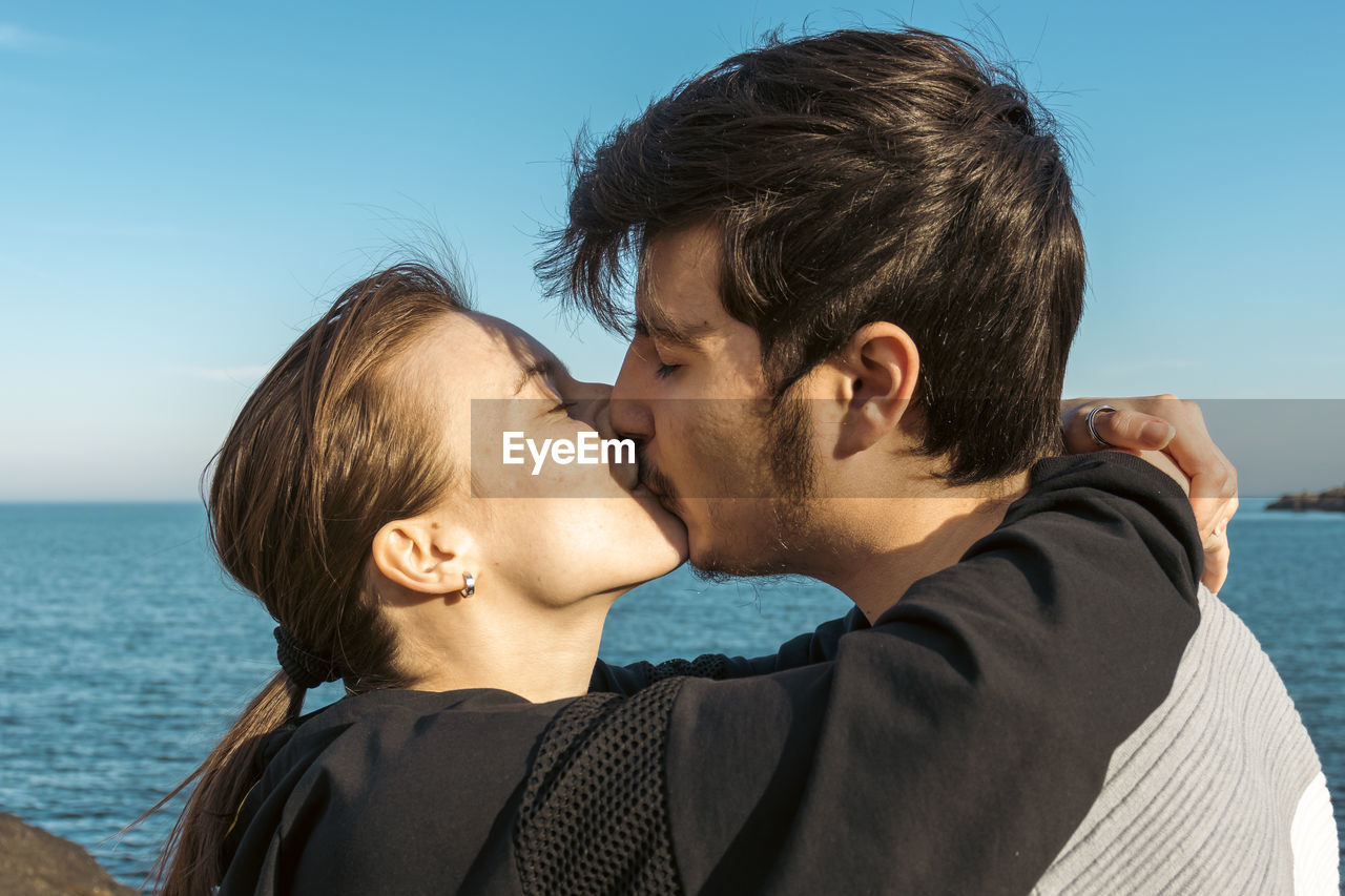 two people, men, kissing, togetherness, love, positive emotion, adult, emotion, water, romance, sea, young adult, sky, portrait, nature, embracing, happiness, women, headshot, bonding, affectionate, beach, person, smiling, land, vacation, blue, leisure activity, trip, ceremony, holiday, casual clothing, lifestyles, day, clear sky, female, outdoors, brown hair, enjoyment, cheerful, sunlight, summer, travel, beauty in nature, eyes closed, relaxation