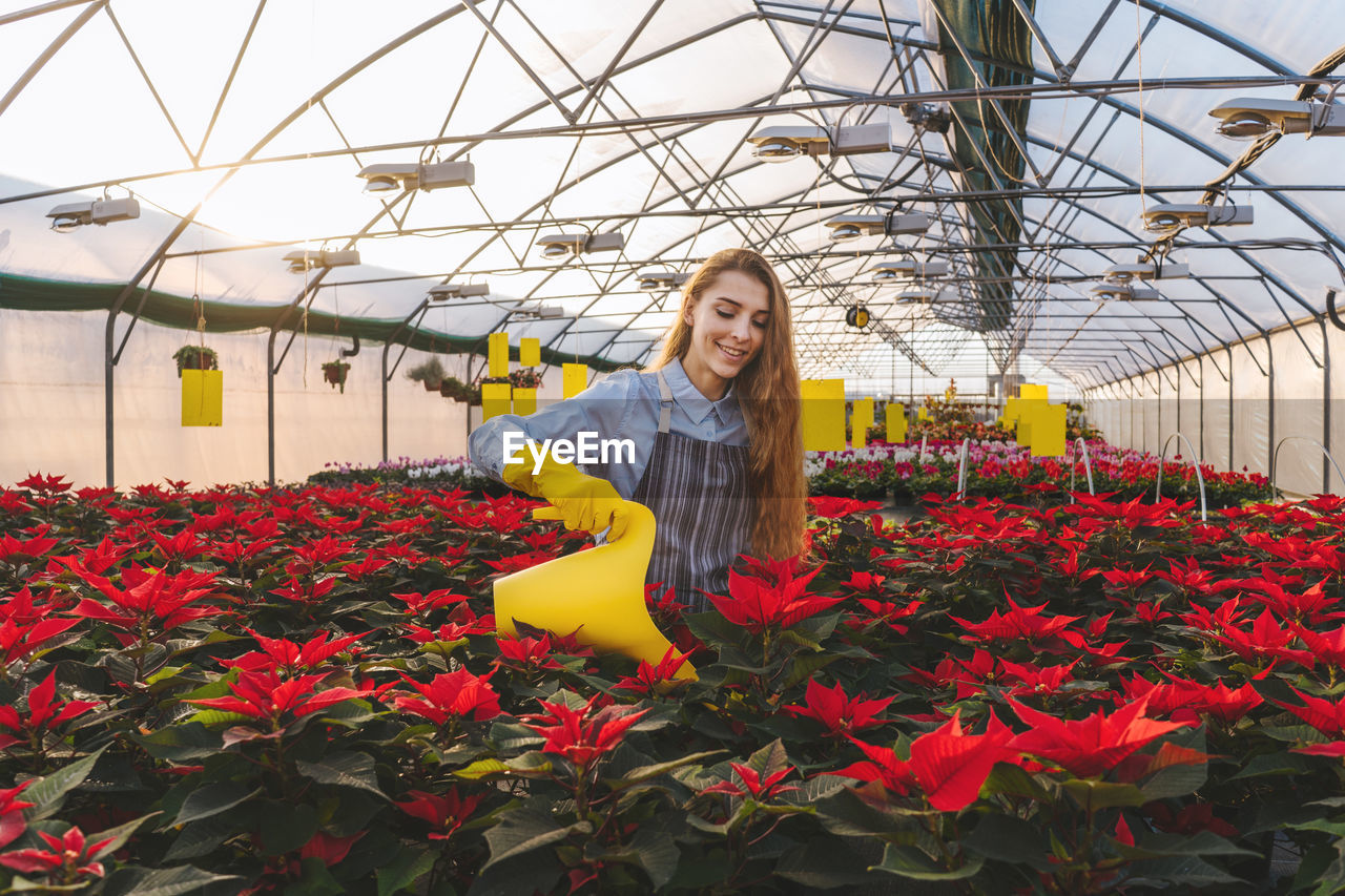 Smiling young woman watering plants while standing in greenhouse