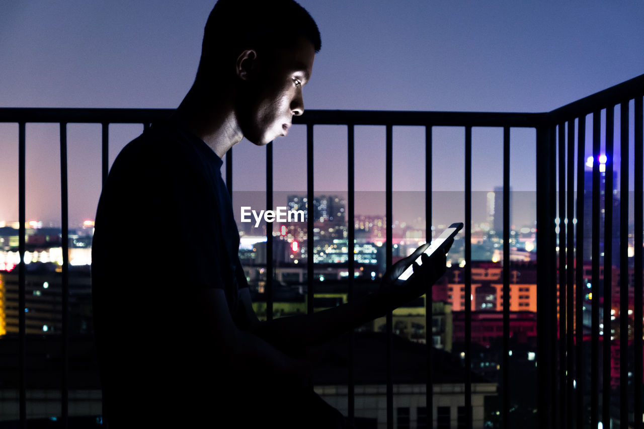 Side view of man using mobile phone while sitting in balcony against city at night