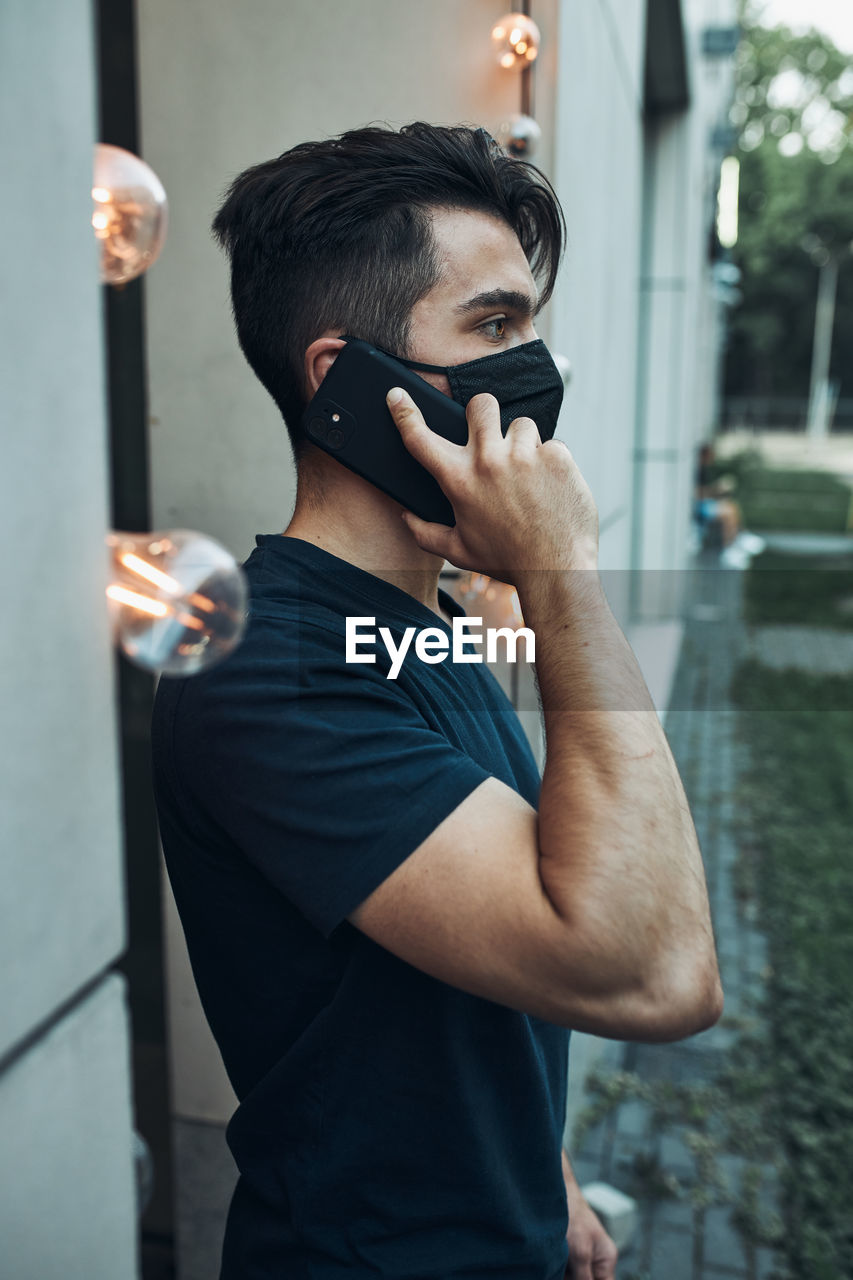 Young man standing in front of restaurant talking on phone using smartphone wearing the face mask