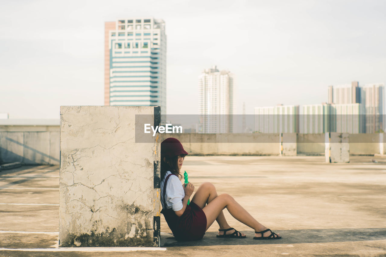 WOMAN SITTING ON MODERN BUILDING AGAINST SKY