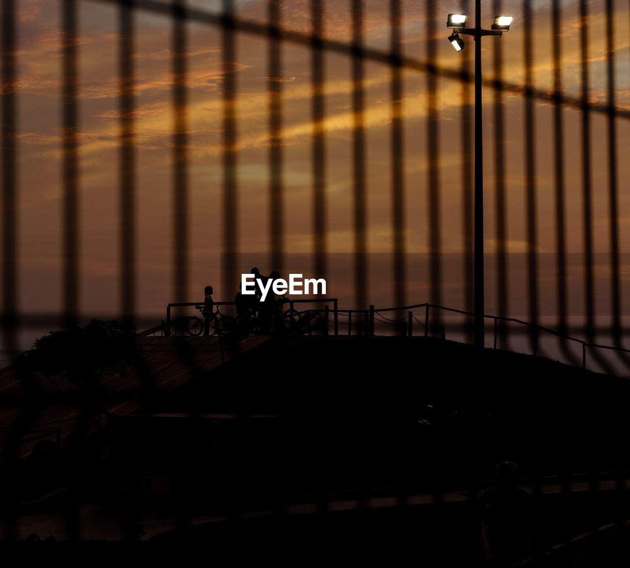 Silhouette family at skateboard park seen through fence against sky during sunset