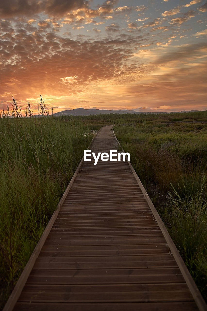 Boardwalk leading towards field against sky during sunset