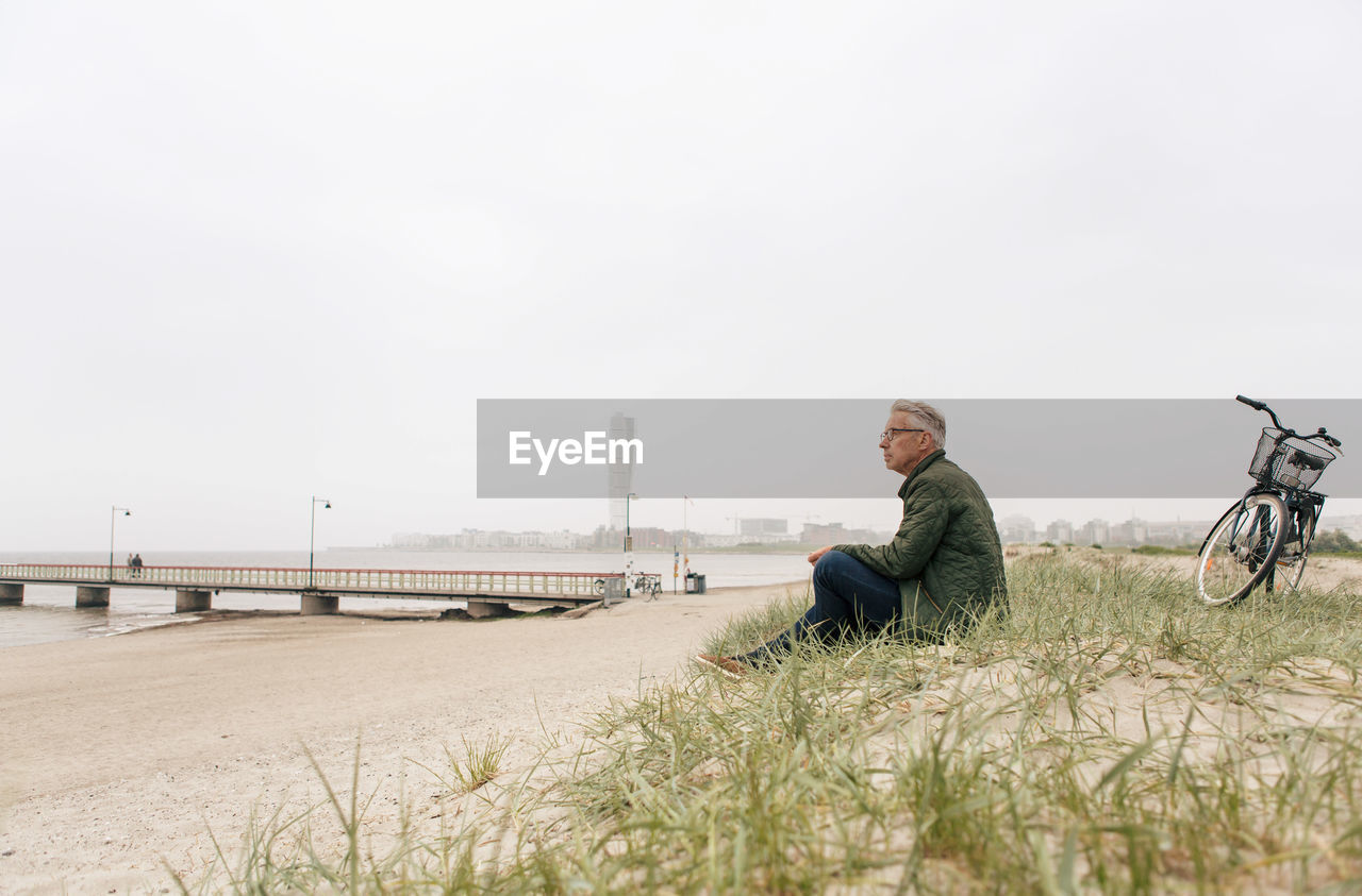 Full length of senior male commuter sitting on sand at beach against clear sky