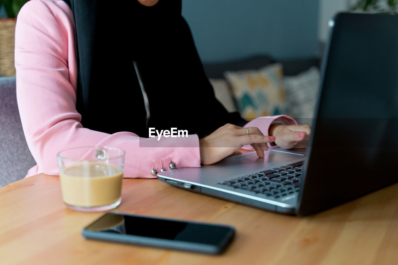 MIDSECTION OF WOMAN USING LAPTOP ON TABLE WITH MOBILE PHONE