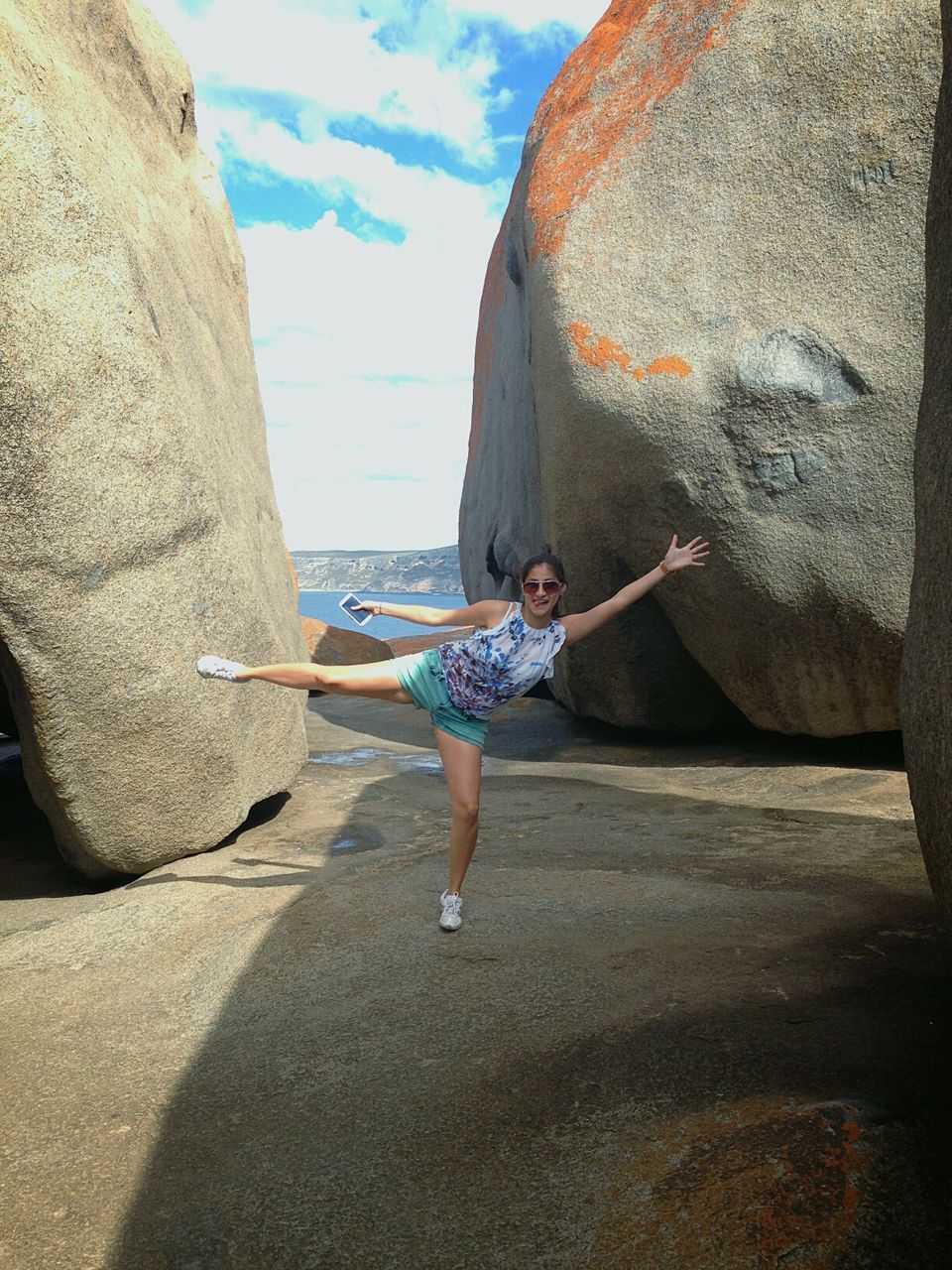 Playful woman standing on one leg over rock formation against sky during sunny day