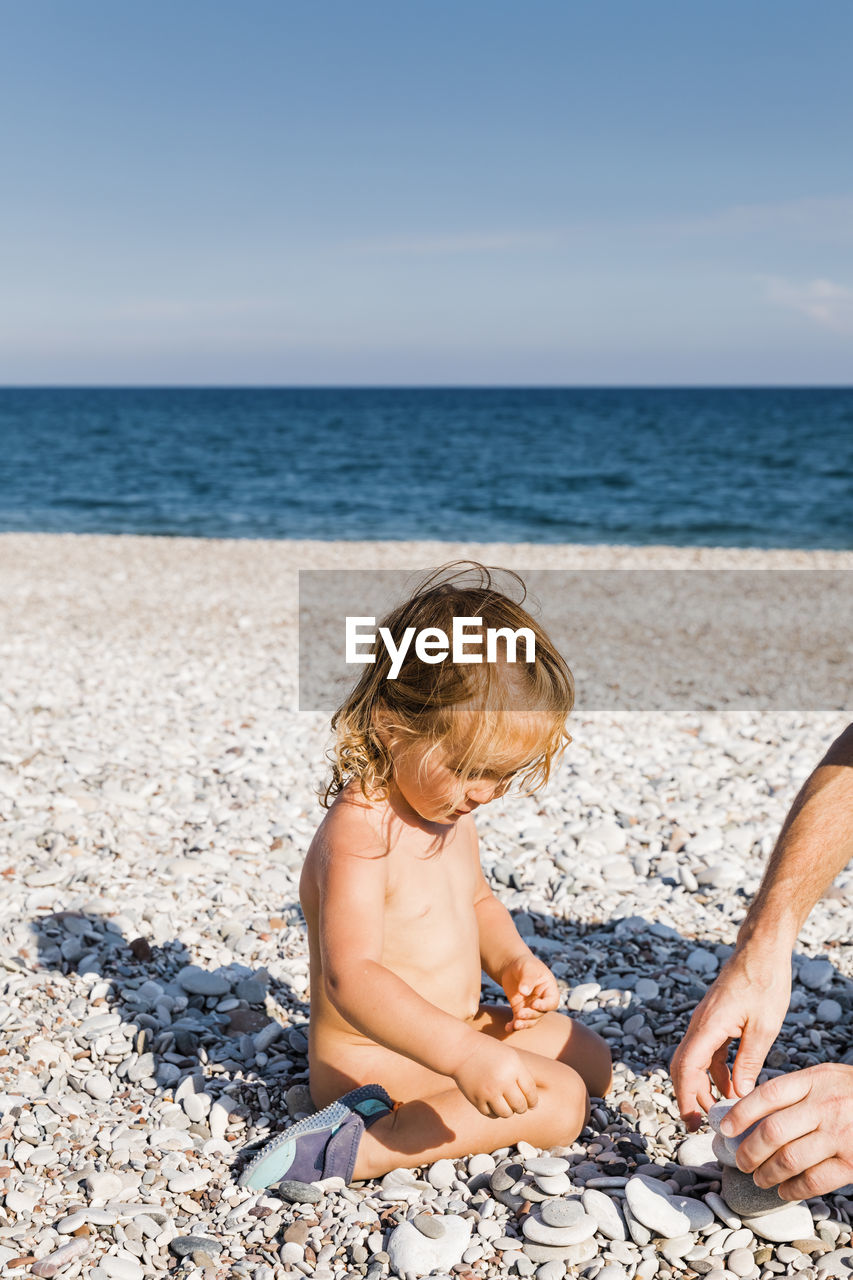 Full length of cute naked girl playing with pebbles at beach during sunny day