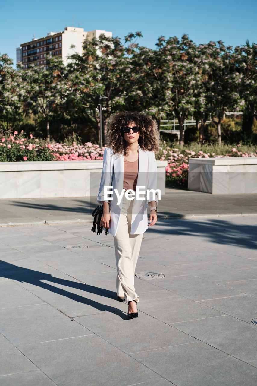 Full body hispanic female manager in trendy white suit and sunglasses with curly hair looking away while walking on the street on sunny day in city