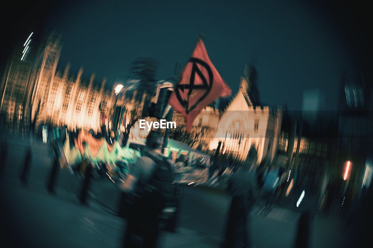 BLURRED MOTION OF PEOPLE ON ILLUMINATED STREET AMIDST BUILDINGS IN CITY